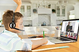 Caucasian boy raising hand for video call, with smiling diverse elementary school pupils on screen