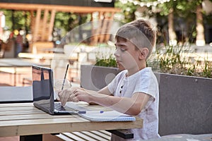 caucasian boy sitting at veranda of cafe, studying with laptop on sunny day