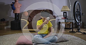 Caucasian boy in VR googles holding remote control and bending. Schoolboy in yellow T-shirt and blue jeans using