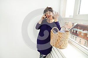 Caucasian brunette woman emotionally, gesturing, talking on phone . dirty laundry basket with clothes. homework concept