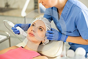 Caucasian brunette young girl lying in spa while having facial treatment procedures with modern technologies vibrating