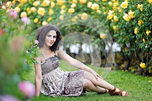 Caucasian brunette young woman sitting on green grass in a rose garden near yellow roses bush, smiling with teeth, looking to the