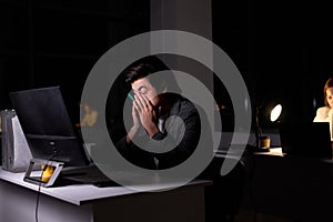 caucasian brunnete man in suit sits at night in office, eyes hurt from overexertion