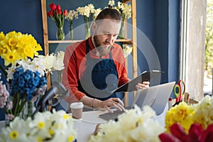 Caucasian businessman in apron in small floral shop using laptop computer and clipboard for order details. Male florist