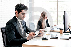 The Caucasian businessman jots something in a small notebook while looking at a computer monitor at his desk workplace.