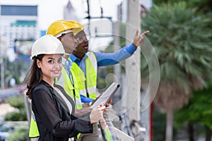 caucasian businesswoman in black suit wear helmet, holding tablet, looking at camera while black engineer colleauges working at photo