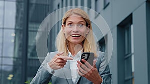 Caucasian businesswoman holding phone stands outdoors enthusiastic happy woman pointing at smartphone screen getting