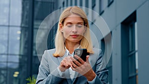 Caucasian businesswoman holding phone stands outdoors enthusiastic happy woman pointing at smartphone screen getting