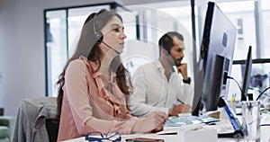 Caucasian businesswoman sitting at desk talking using phone headset and computer in busy office