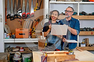 Caucasian carpenter worker man stand with his Asian wife who hold laptop that work with wood working in factory workplace and they