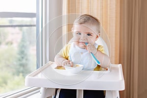Caucasian child boy with dirty messy face sitting in high chair eating apple puree with spoon