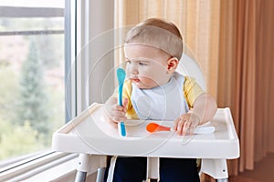 Caucasian child boy with dirty messy face sitting in high chair eating apple puree with spoon