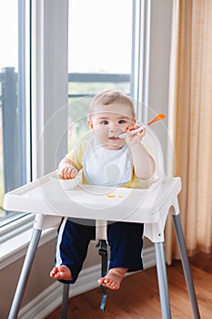 Caucasian child boy with dirty messy face sitting in high chair eating apple puree with spoon