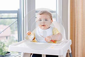 Caucasian child boy with dirty messy face sitting in high chair eating apple puree with spoon