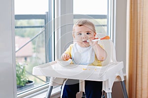 Caucasian child boy with dirty messy face sitting in high chair eating apple puree with spoon