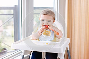 Caucasian child boy with dirty messy face sitting in high chair eating apple puree with spoon