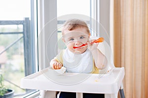 Caucasian child boy with dirty messy face sitting in high chair eating apple puree with spoon