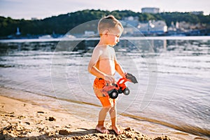 Caucasian child boy playing toy red tractor, excavator on a sandy beach by the river in red shorts at sunset day