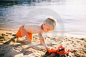 Caucasian child boy playing toy red tractor, excavator on a sandy beach by the river in red shorts at sunset day