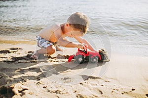 Caucasian child boy plays toy red tractor, excavator on sandy beach by the river in shorts at sunset day