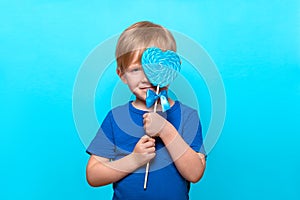 Caucasian child hold huge heart shaped lollipop on stick, close his eyes and smile. Studio shot with copy space on blue solid