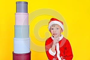 Caucasian child sits near a tower of round gift boxes, holds a finger to his mouth, urging with this gesture that it is a secret