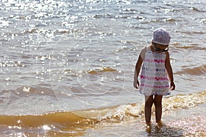 Caucasian child of three years in sunglasses standing on the river beach washed by waves looking down