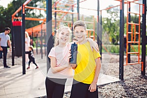 Caucasian children brother and sister stand embrace at outdoor sports ground with smile and flask with water after training. Twins