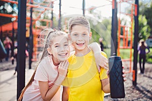 Caucasian children brother and sister stand embrace at outdoor sports ground with smile and flask with water after training. Twins