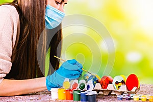 Caucasian concentrated girl in medical mask and disposable gloves decorates handmade eggs for the pascal holidays in