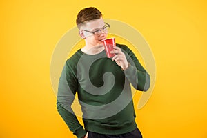 Portrait of young handsome confident guy in green sweater holding red plastic cup on yellow background