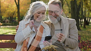 Caucasian couple happy 60s old mature woman man hold credit card mobile phone bank app two grandparents in city park