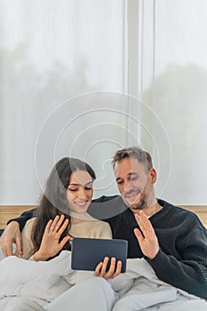 Caucasian couple having good time using tablet computer making video call together on bed in bedroom at home