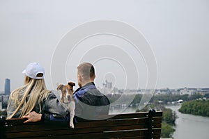 Caucasian couple in love sitting on wooden bench with a jack Russell terrier puppy and enjoying cityscape of Belgrade