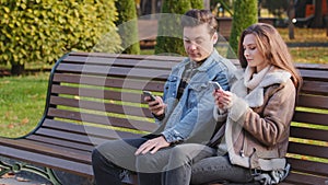 Caucasian couple two young people partners sit on bench use phone cell gadgets scroll internet web pages check social