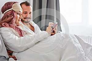 Caucasian couple using smartphone lying on bed together, checking social networks news