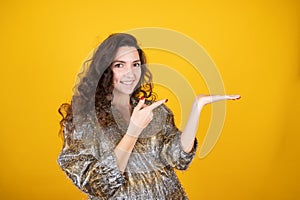 Curly woman pretending to hold something on the palm pointing with a finger isolated on yellow studio background