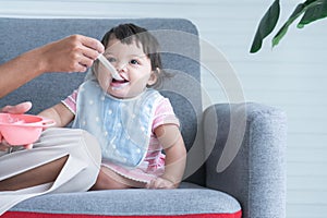 Caucasian Cute 7 month newborn baby girl eating blend mashed food, sitting on sofa with apron at home, mother hand is holding