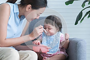 Caucasian Cute 7 month newborn baby girl eating blend mashed food, sitting on sofa with apron at home, young Asian mother holding