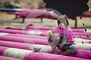 Caucasian cute boy sits on a pink pipes with black paper boats and clouds at the construction site