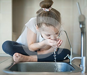 Caucasian cute girl is drinking from water tap or faucet in kitchen at home. Hydration. Lifestyle. Healthcare, healthy drink