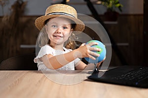 Caucasian cute little girl in a hat sits alone in the evening at a table with a laptop holding a globe in her hands.