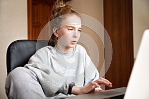 Caucasian cute red-haired teenage girl in a grey hoodie and pants, sitting at a table in front of a laptop indoors. Home