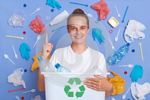 Caucasian eco-friendly woman holding box full of a plastic bottles for recycling, standing surrounded with much garbage, pointing