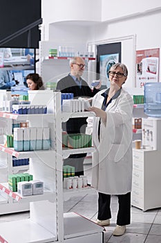 Caucasian elderly pharmacist standing in drugstore arranging shelves while checking pills packages
