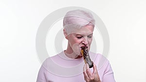 Caucasian elderly woman bites an eclair with pleasure over white background