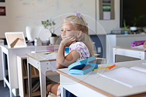 Caucasian elementary schoolgirl with hand on chin looking away while sitting at desk in classroom