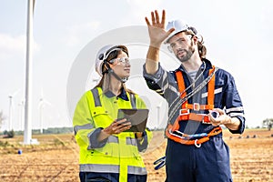 Caucasian engineer man discuss together with co-worker woman hold blueprint with tablet stay in front of row of windmill