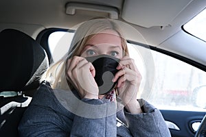 Caucasian european woman sitting in a car and wearing black surgical medical face mask as a way of protection against coronavirus