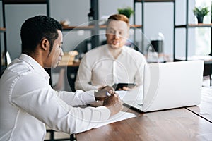 Caucasian European young businessman explaining to African coworker business plan sitting at desk